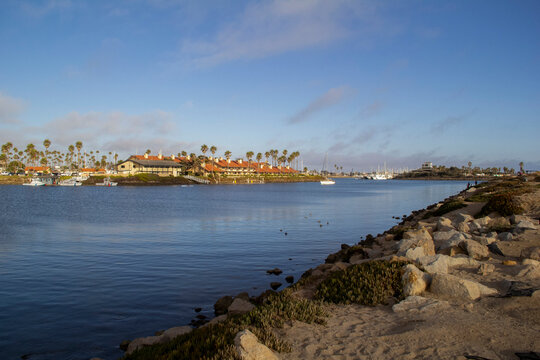Rocky shoreline next to marina with ships and buildings.