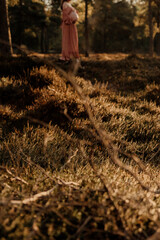 Pregnant woman in pink dress standing in forest meadow at sunset.