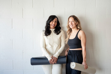 Two women smile with yoga mats against a studio wall