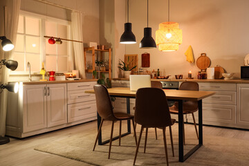Interior of modern kitchen with glowing lamps, white counter, chairs and laptop on dining table at evening