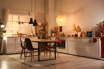 Interior of modern kitchen with glowing lamps, white counter, chairs and laptop on dining table at evening
