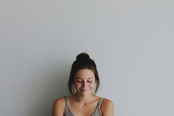 Fototapeta premium Shy young woman looking down and smiling gently with a bun hairdo against a simple gray wall