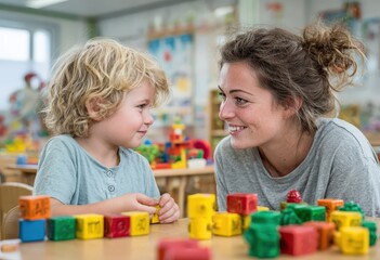 Young woman engaging with a child, both smiling while playing with colorful building blocks on a wooden table in a bright, cheerful classroom environment, fostering creativity and learning