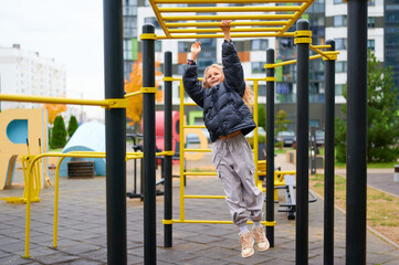 child playing outdoors on monkey bars at urban playground, displaying joy and physical activity. young girl dressed warmly, enjoying recreational time. modern cityscape backdrop creates vibrant scene © khmelev