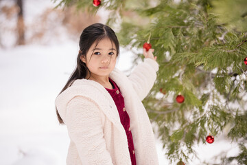 Girl in fleece coat decorates pine tree with red ornaments in snow.
