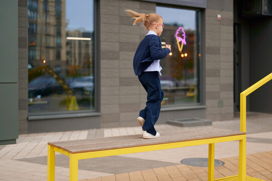 girl in school uniform enjoying playful outdoor activity, balancing on yellow bench near modern building. captures essence of childhood fun and urban lifestyle balance