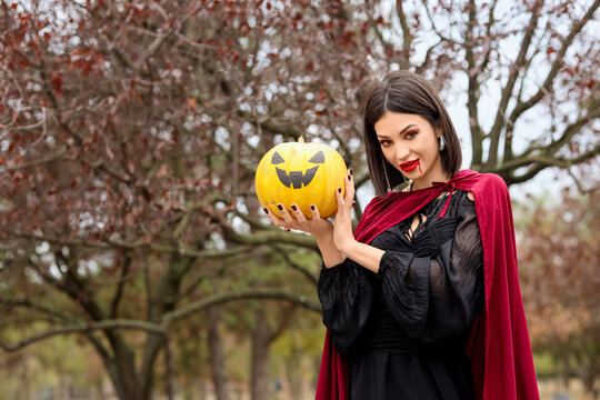 Young woman in vampire costume holding jack-o'-lantern pumpkin for halloween celebration and autumn holiday festivities, the woman wears a black dress and a red cape