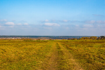 Vast autumn field with a clear dirt road stretching into the distance, framed by a distant forest and a quaint village under a bright, expansive blue sky