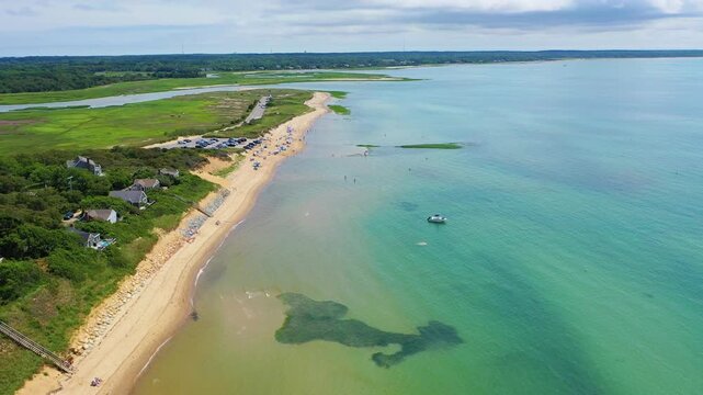 Drone pass along a shoreline reveals swimmers in shallow surf, kayaks drifting nearby, and rows of coastal homes nestled in trees overlooking sandy beaches and clear turquoise waters.