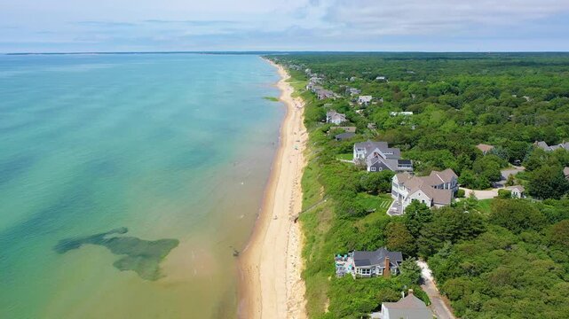 Maine beach scene with families wading in turquoise water, kayaks drifting nearby, and traditional coastal cottages dotting the shoreline framed by lush green trees and open sandy beaches.