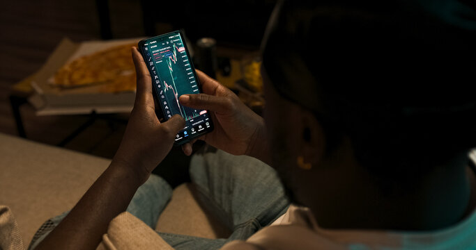 A trader observes financial indicators on his phone and laptop. Cryptocurrency chart movements and market analysis in progress. Shot from behind.