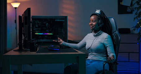 A professional female player enjoys a team victory during an online match. She jokes with friends via headset while sitting in a dark room illuminated by soft blue LEDs.