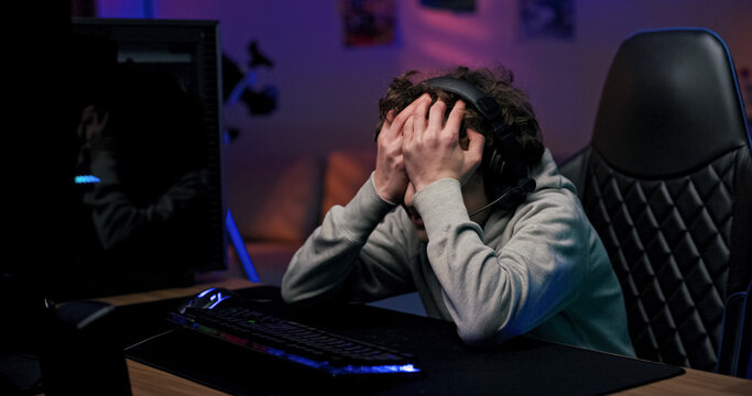 A young gamer experiences disappointment after a video game defeat. The child wears a headset, rests his head on his hand, and stays in touch with teammates online.