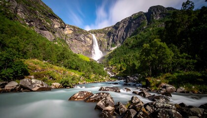 A breathtaking view of a mountain waterfall cascading into a flowing river, embraced by lush green cliffs under a bright blue sky