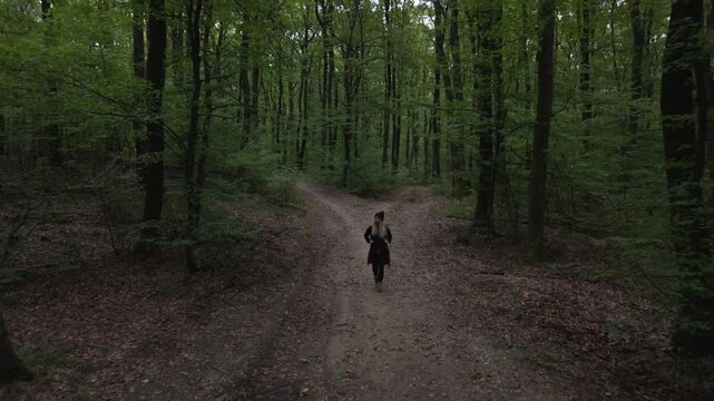 A woman walks alone on a dirt path in a dense forest, approaching a fork in the road. The scene captures a quiet moment of decision and exploration surrounded by green trees and natural beauty.