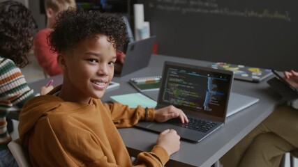 Medium portrait of happy 12-year-old polyethnic African American boy in hoodie sitting at desk at lesson, working on computer code on laptop, looking at camera and smiling confidently - Powered by Adobe