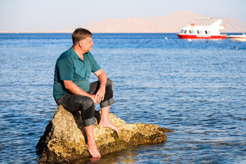 A blond man in clothes sits on a rock in the sea far from the shore.