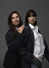 Two business women in blazers and jeans posing on a white cyclorama, sitting on the floor or chair with a constant light behind them. One woman puts her hand in the other's pocket. Minimalist photos