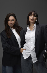 Two business women in blazers and jeans posing on a white cyclorama, sitting on the floor or chair with a constant light behind them. One woman puts her hand in the other's pocket. Minimalist photos