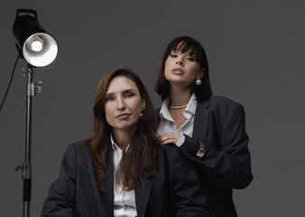 Two business women in blazers and jeans posing on a white cyclorama, sitting on the floor or chair with a constant light behind them. One woman puts her hand in the other's pocket. Minimalist photos