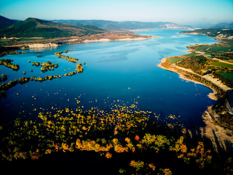 Aerial view of a tranquil blue reservoir reflecting the sky, surrounded by lush green hills and vibrant autumn foliage, Mediano, Arag&Atilde;&sup3;n, Spain.