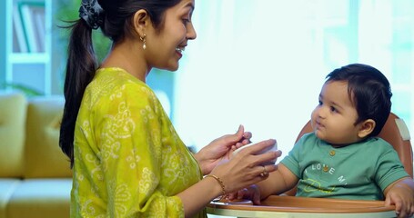Indian infant eating baby food, mother feeding with spoon while sitting in modern living room, showing love, care, healthy nutrition, bonding, and early parenting moments focused on child’s growth - Powered by Adobe