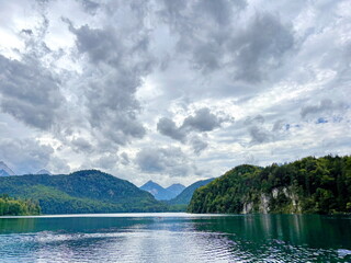 Amazing panorama of the calm emerald surface of the Alpsee mountain lake reflecting the clouds hanging over it surrounded by green forest hills.