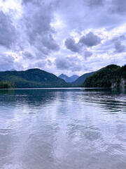 Amazing panorama of the calm emerald surface of the Alpsee mountain lake reflecting the clouds hanging over it surrounded by green forest hills.
