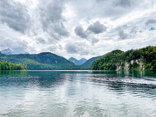 Amazing panorama of the calm emerald surface of the Alpsee mountain lake reflecting the clouds hanging over it surrounded by green forest hills.