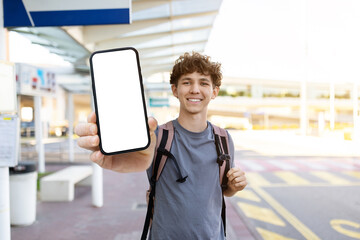 Young man smiling and showing smartphone with blank mockup screen at airport. Concept of app, travel ticket, or online booking