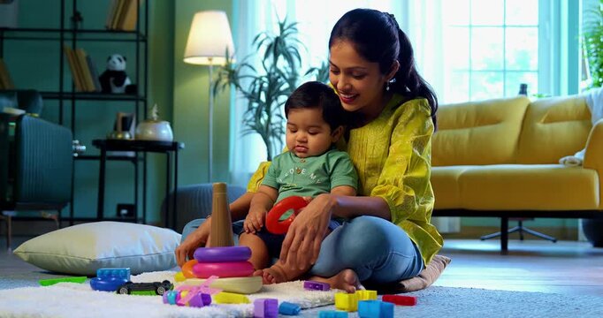Indian infant boy playing ring toys with young mother while sitting on carpet in modern living room, sharing joyful moments, love, affection, bonding, and happiness of parenthood