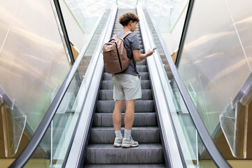Rear view of young traveler guy standing on escalator at airport, holding smartphone and backpack. Modern transport and travel concept, full length