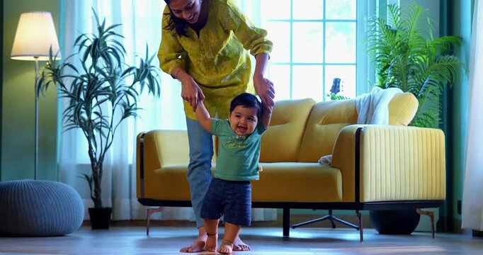 Indian infant boy walking holding young mother's hand in modern living room, learning to balance and take first steps with love, care, affection, and gentle guidance from mom