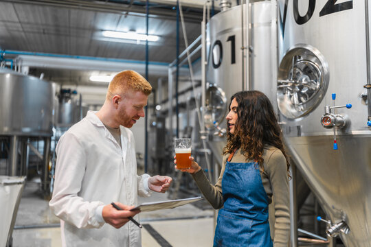 Two brewers in work uniforms inspect and taste craft beer beside stainless steel fermentation tanks, discussing quality control and process