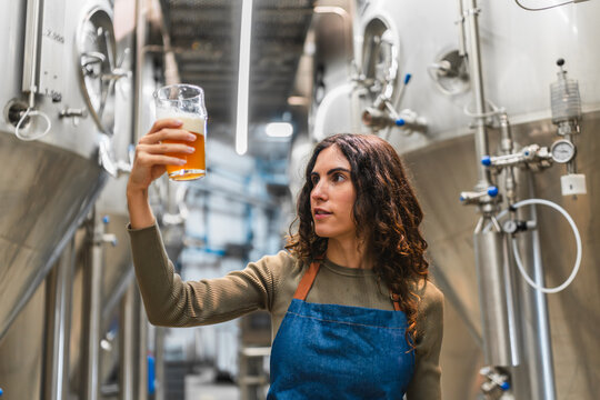 Female brewer holding a glass of beer, examining clarity and color during quality control in a modern craft beer facility