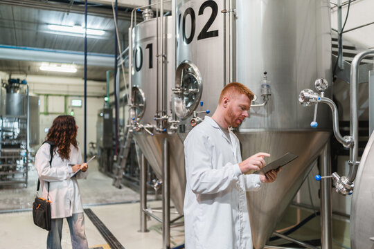 Brewery workers in lab coats inspecting stainless steel fermentation tanks, ensuring quality control in the beverage industry