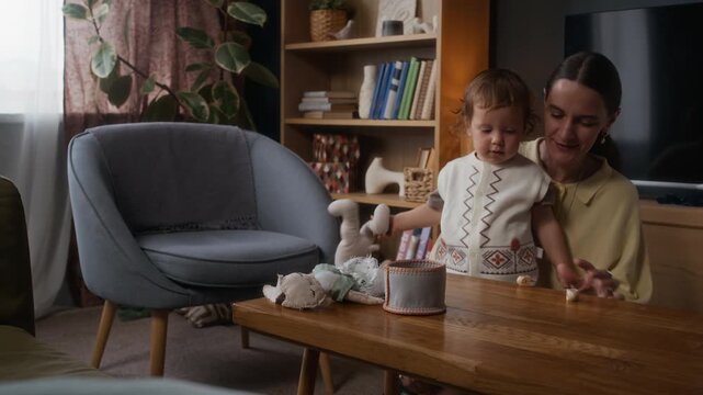Medium long shot of young Jewish woman sitting at table with her playful toddler son in modern cozy apartment and showing him how to spin dreidels at Hanukkah