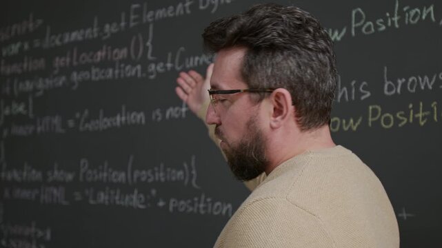 Medium close-up of nerdy young Caucasian male programming teacher conducting lesson, explaining coding algorithm, pointing at variables and commands, typing example on tablet