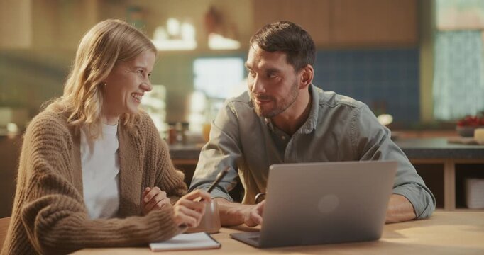 Domestic Teamwork Moment at the Kitchen Table. Woman Takes Notes in a Notebook while Her Husband Shows Her the Laptop Screen, Discussing Home Renovating Ideas or Travel Destinations Over Coffee