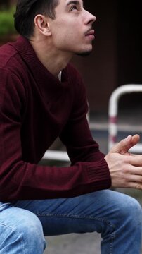 Vertical: A young man with dark hair in a burgundy sweater sits outdoors showing despair and emotional vulnerability, expressing frustration and contemplation in an urban environment.