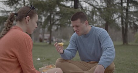 A young couple shares a funny moment during their outdoor picnic in the park. The man makes a sour face while eating fruit, bringing laughter and joy to their special date.