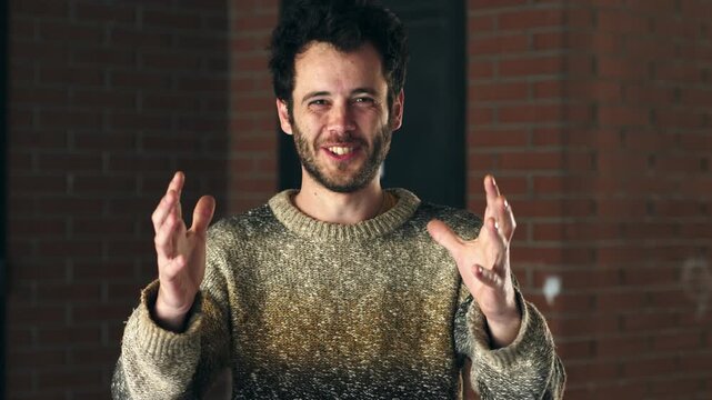 A young man with dark hair in a sweater laughs while gesturing with his hands to indicate something very big, showing humor, exaggeration, and playful energy indoors.
