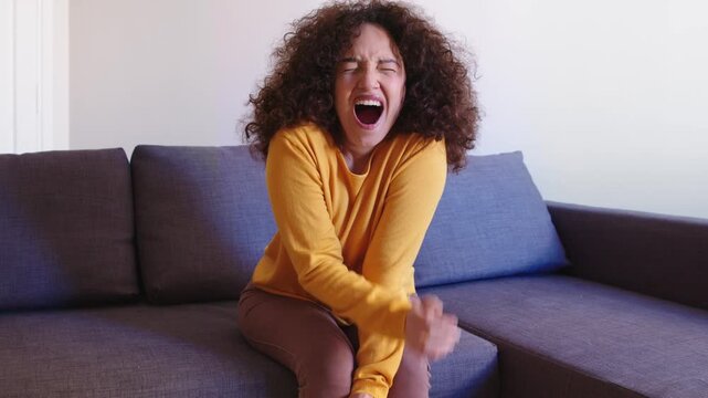 A young South American woman with Latin features yawns on the sofa while watching TV, showing boredom, fatigue, and casual home relaxation.