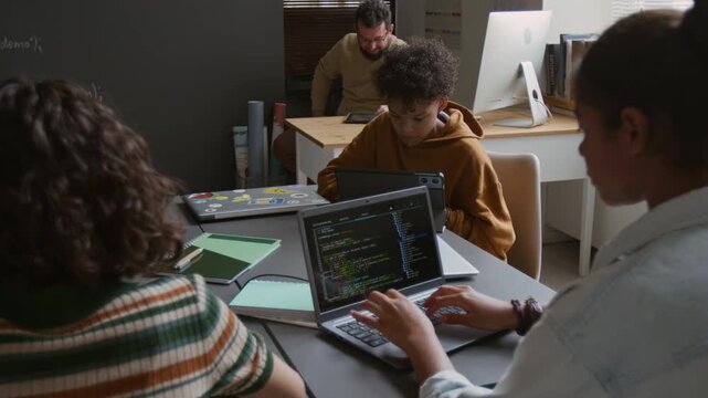 Medium shot of busy diverse male and female students in casual clothes sitting around table at information technology class, working on coding tasks on laptops, teacher observing from corner desk