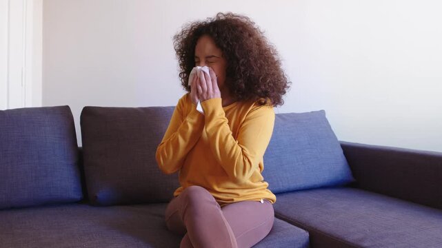 A young Latin woman in a yellow sweater blows her nose on the sofa, struggling with an allergy attack, showing discomfort and a vulnerable moment at home.