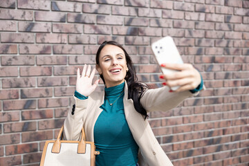 A joyful woman waves at her selfie while posing confidently outdoors, radiating happiness and energy in a chic outfit.