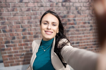 A stylish woman smiles brightly for a selfie against a detailed brick wall, showcasing joy and self-assurance.