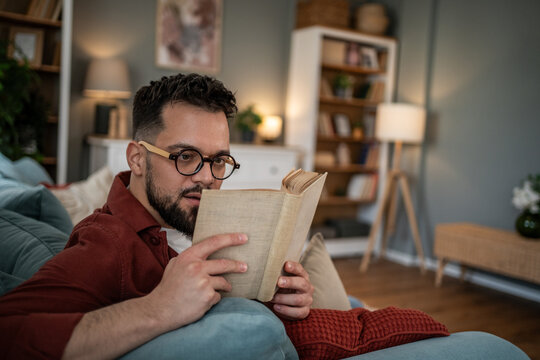 Young man relaxing at home reading book with glasses - Powered by Adobe