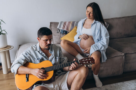 Couple at home with pregnant woman relaxing as partner plays guitar