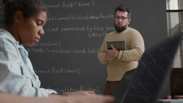 Medium shot of geeky young Caucasian male IT teacher standing by blackboard, explaining coding algorithm, pointing at variables and structure during lesson, girls working on laptops in foreground